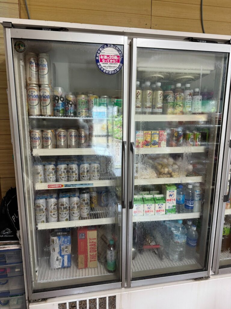 冷蔵庫に並ぶオリオンビール缶、酎ハイ、ソフトドリンク、そしてマグロの刺身パック/Fridge stocked with Orion beer, chu-hi, soft drinks, and fresh tuna sashimi